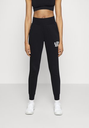 Persoon draagt zwarte high-waist joggers met wit "Björn Borg" logo op de dij, zwarte crop top, witte sokken en witte sneakers, rechtopstaand.