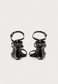Sandales à talons en cuir verni noir avec brides de cheville, design à bout ouvert et un talon bloc étroit, présentant un détail logo métallique à l'arrière.
