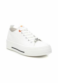Zapatillas de lona blancas con una suela gruesa y texturizada, banda de acento negra circular, detalles en ojetes naranjas y hardware plateado en los cordones.