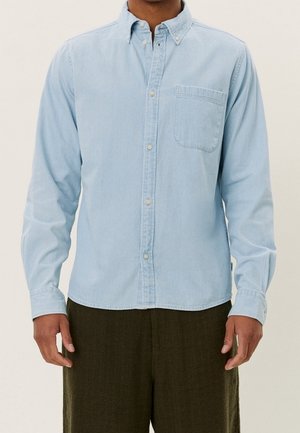 Mand iført lyseblå skjorte med brystlomme og mørkebrune bukser, stående foran en neutral baggrund.