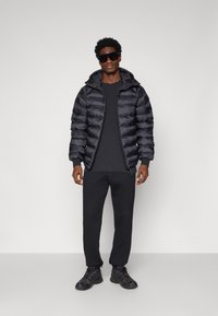 Giacca a vento nera con cappuccio, caratterizzata da un design trapuntato. Indossata sopra una maglietta nera a maniche lunghe e abbinata a pantaloni della tuta neri e sneakers.