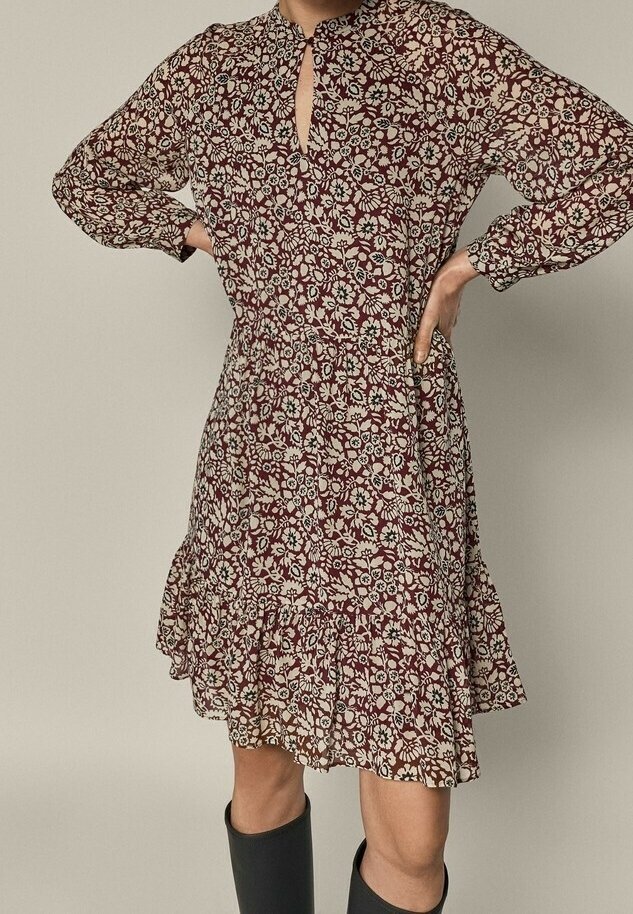 Femme portant une robe marron fleurie avec des manches longues et un ourlet à volants, les mains sur les hanches, associée à des bottes noires montantes jusqu'aux genoux.