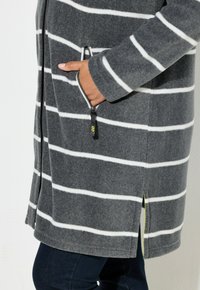 Cappotto a righe grigie con linee bianche, realizzato in tessuto morbido. Presenta una cerniera frontale, tasche laterali e uno spacco laterale che rivela un dettaglio giallo.