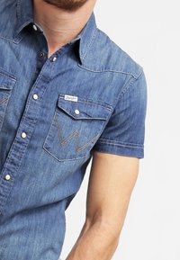 Ljusblå jeansskjorta med kort ärm, utrustad med två bröstfickor, vit sömnad och en märkesetikett ovanför den vänstra fickan.