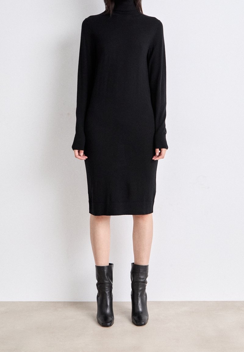 Osoba ubrana w czarną, długą sukienkę z golfem i czarne skórzane botki do połowy łydki, stojąca na beżowej podłodze.