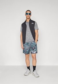 Hombre con gafas de sol reflectantes azules, camisa gris de manga corta, chaleco negro sin mangas, pantalones cortos estampados, calcetines negros y zapatillas blancas.