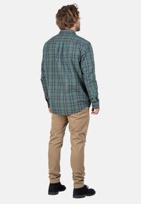 Mann mit braunen Haaren, der ein langärmliges grünkariertes Hemd, beigen Hosen und schwarze Schuhe trägt. Er steht mit dem Rücken zu einem weißen Hintergrund.