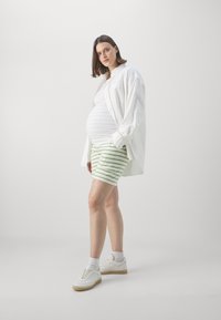 Graviditetsantrekk som består av en hvit skjorte med knapper, en tettsittende hvit topp og grønnstripete shorts. Modellen har på seg hvite joggesko og ankelstrømper.