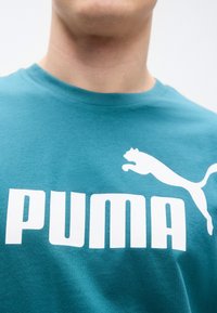 Τιρκουάζ βαμβακερό T-shirt με λευκό λογότυπο "PUMA" και εικονίδιο αλματώδους πάνθηρα. Στρογγυλή λαιμόκοψη και κοντά μανίκια. Ομαλή υφή.