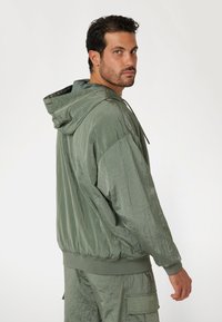 Zöld kapucnis dzseki fényes anyagból, laza szabású, rugalmas mandzsettákkal és elülső kenguru zsebbel. Tökéletesen párosítva hasonló anyagú nadrággal.