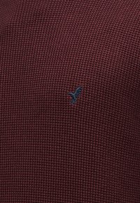 Pull bordeaux texturé présentant un petit logo brodé d'un oiseau bleu marine sur le côté gauche. Le matériau a un motif tissé.