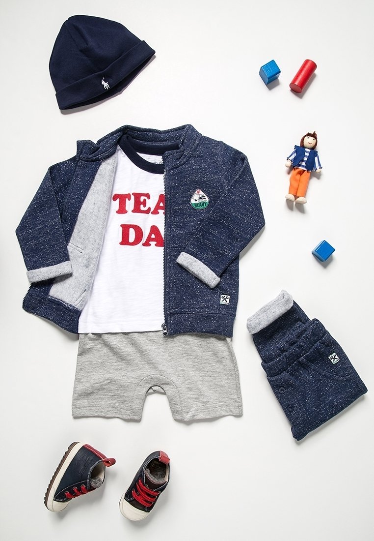 Veste et casquette marine, t-shirt blanc « TEAM DAD », short gris, pantalon marine, chaussures de bébé, poupée et trois blocs en bois sur fond blanc.