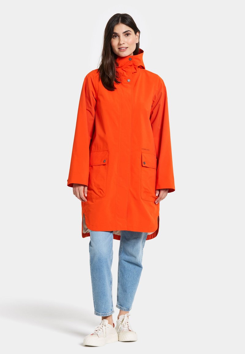 Orangen wasserfester Regenmantel mit hohem Kragen, zwei Vordertaschen, seitlichen Schlitzen und einer lockeren Passform, kombiniert mit hellblauen Jeans und weißen Sneakers.