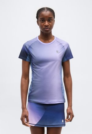 Žena v modro-fialovom prechádzajúcom športovom krátkom rukáve a zodpovedajúcej sukni stojaca na jednoduchom svetlom pozadí.