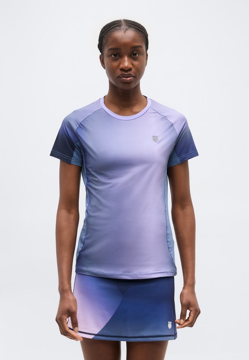 Kvinde iført en kortærmet atletisk skjorte med blå og lilla gradient samt matchende nederdel, stående mod en ensfarvet lys baggrund.