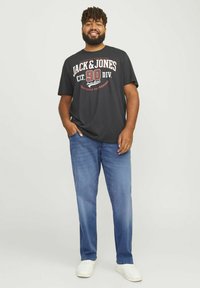 T-shirt nera con grafica testuale rossa e bianca, abbinata a jeans di denim blu e sneakers bianche, in piedi con le mani sui fianchi su uno sfondo chiaro.