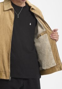 Veste en corduroy beige avec un intérieur en polaire, fermeture éclair à l'avant, et une chemise noire avec un petit logo blanc en dessous.