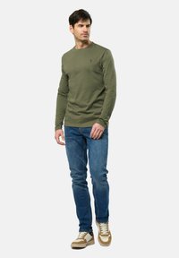 Haut à manches longues vert olive avec un col rond et un logo brodé, associé à un jean bleu et des baskets beiges. Coupe décontractée et tissu.