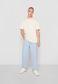 Cream cotton t-shirt with short sleeves and round neck, paired with light blue baggy jeans and white sneakers. Simple, casual outfit.