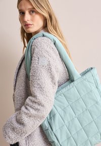 Wypchana torba w kolorze miętowym o pikowanej fakturze, z krótkimi uchwytami oraz szara kurtka polarowa z naszywką na ramieniu.