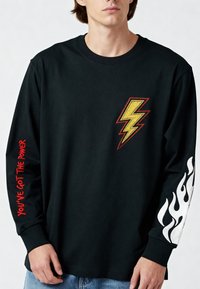 Persoon draagt een zwart shirt met lange mouwen en een gele bliksemschicht op de borst, rode tekst "YOU'VE GOT THE POWER" op de linker mouw, en een wit vlammenontwerp op de rechter mouw.