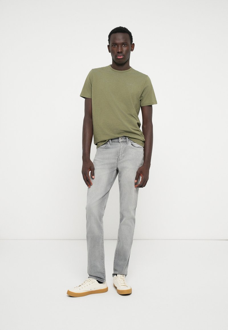 Zelené bavlněné tričko, šedé džíny a béžové tenisky. Model stojí vzpřímeně na bílém pozadí a předvádí ležérní outfit.