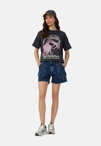 T-shirt graphique gris foncé avec des accents roses et le logo du groupe, associé à un short en denim cargo. Complété par une casquette grise et des chaussures de sport.