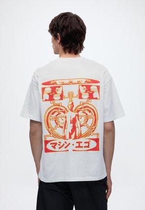 Homme aux cheveux foncés portant un t-shirt blanc avec un graphisme futuriste orange et rouge représentant une figure debout et du texte japonais dans le dos.