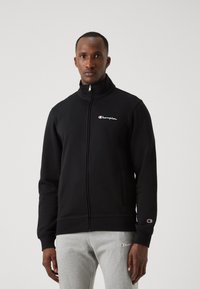 Sudadera con cremallera negra, hecha de tejido suave, que cuenta con un alto cuello, bolsillos frontales y un pequeño logo de Champion en el pecho y en la manga.