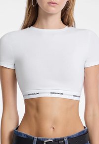 Witte korte mouwen crop top gemaakt van zachte stof, met een zwart merkembleem elastische band aan de onderkant met de tekst "GUESS JEANS".