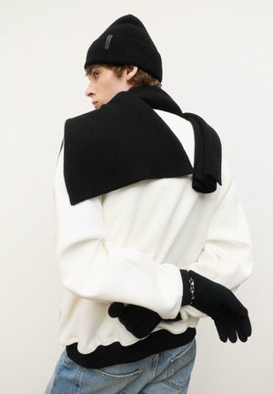 Beanie tricotat negru, eșarfă și mănuși purtate cu un hanorac alb, având o textură cu nervuri și un design casual. Mâinile sunt împletite în spate.