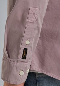 Camicetta a maniche lunghe rosa chiaro con polsini a bottone, caratterizzata da una toppa ricamata e un'etichetta con logo. Tessuto di cotone liscio e bottoni bianchi.