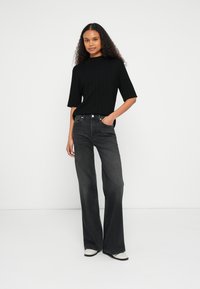 Top negro de punto acanalado con mangas cortas, combinado con vaqueros acampanados de tiro alto en gris oscuro. La modelo posa con confianza, con una mano en el bolsillo.