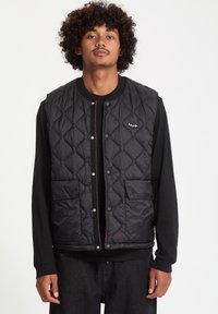 Quiltet sort vest med trykknapper, to frontlommer, kontrastsyninger og et logo-patch på brystet; lagd oven på en sort langærmet skjorte.