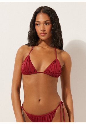 Vrouw met golvend donker haar, dragend een diep rood gerimpeld driehoekig bikinibovenstuk en bijpassende bikinitruisjes met zijstrikjes, tegen een neutrale achtergrond.