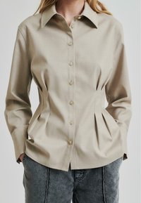 Beige skjorte med knapper, strukturert design, lange ermer og sidelister, med lysebrune knapper og spiss krage.
