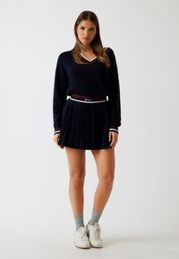 Marineblauer Pullover mit V-Ausschnitt und gestreiften Akzenten, kombiniert mit einem plissierten marineblauen Rock mit einem markierten Taillenbund. Weiße Sneakers und graue Socken.