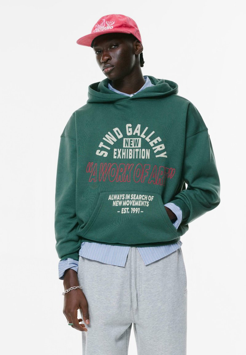 Felpa verde con grafica "STWD GALLERY", pantaloni della tuta grigi sovrapposti a una maglietta a righe blu e un cappellino rosa.