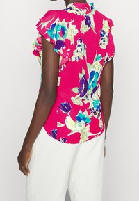 Květinová blůza s volánovými rukávy, s jasně růžovým pozadím a vícebarevnými květinovými vzory v modré, zelené a fialové.