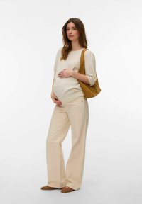 Femme enceinte portant un pull crème et un pantalon, chaussures plates marron, portant un sac épaule en daim beige, tenant son ventre avec les deux mains.