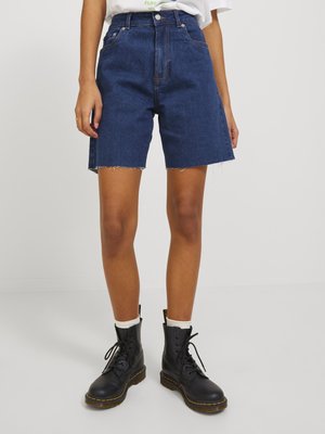 Personne portant un short en jean bleu foncé mi-long avec des bords bruts, des chaussettes blanches et des bottes noires à lacets, debout devant un fond uni.