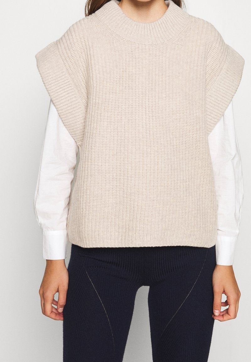 Beige gestrickter Pullover mit hohem Kragen, kurzen Ärmeln und einem gerippten Muster. Getragen über einem weißen Hemd mit strukturierten Manschetten.