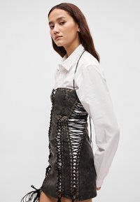 Crna kožna haljina s vezicama sprijeda, metalnim okicama i resama sa strane. Model nosi bijelu košulju s ovratnikom ispod.