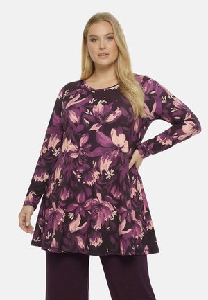 Kvinde iført lilla tunika med stort rosa og violet blomsterprint og matchende mørkelilla bukser, poserer med den ene hånd på hoften.