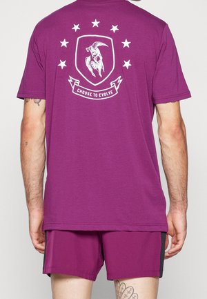 Mann trägt ein lila Hemd und Shorts, auf der Rückseite des Hemdes ist ein Totenkopf-Emblem, sieben Sterne und der Satz „CHOOSE TO EVOLVE.".