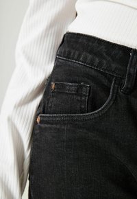 Schwarze Denim-Jeans mit einem klassischen Fünf-Taschen-Design, das ein kleines bronzenes Logo-Element und eine glatte, strukturierte Oberfläche aufweist.