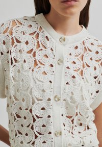 Cardigan scurt de culoare deschisă, cu un model intricate din dantelă, mâneci scurte, guler rotund și închidere cu nasturi.
