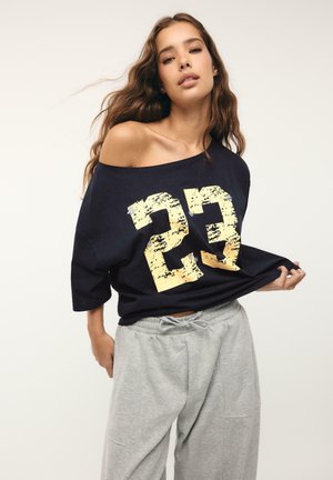 Giovane donna con lunghi capelli ondulati che indossa una camicia nera oversize con il numero giallo "23" e pantaloni della tuta grigio chiaro, in posa su uno sfondo semplice.