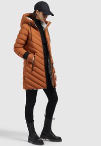 Puffjacka i bränd orange med diagonal sömnad, svarta accenter och huva. Ett par svarta leggings och knähöga svarta stövlar.