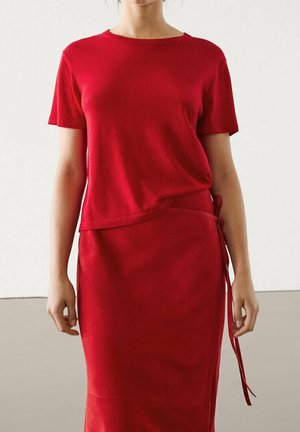 Woman wearing a bright red short-sleeve top and matching skirt with side tie detail, standing against a plain light background.